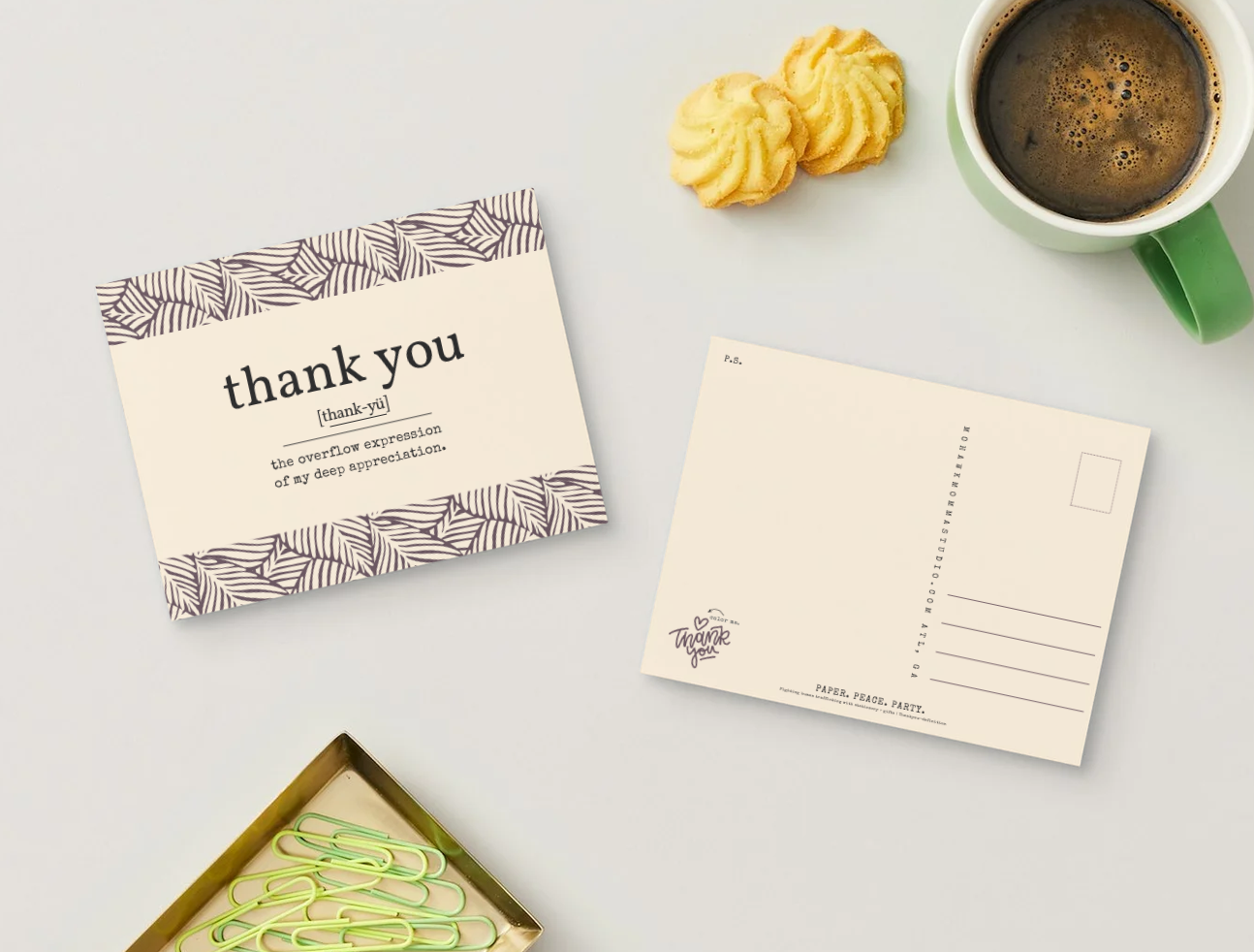 A styled flat-lay featuring a cream-colored 'thank you' postcard with a brown geometric pattern border, alongside an empty postcard, a green coffee mug, two golden pastries, and a tray of paperclips on a light gray surface.