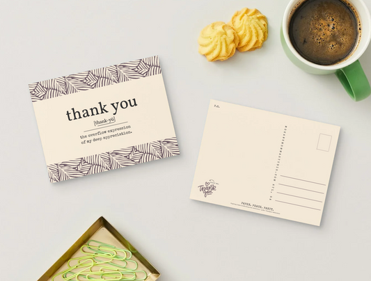 A styled flat-lay featuring a cream-colored 'thank you' postcard with a brown geometric pattern border, alongside an empty postcard, a green coffee mug, two golden pastries, and a tray of paperclips on a light gray surface.