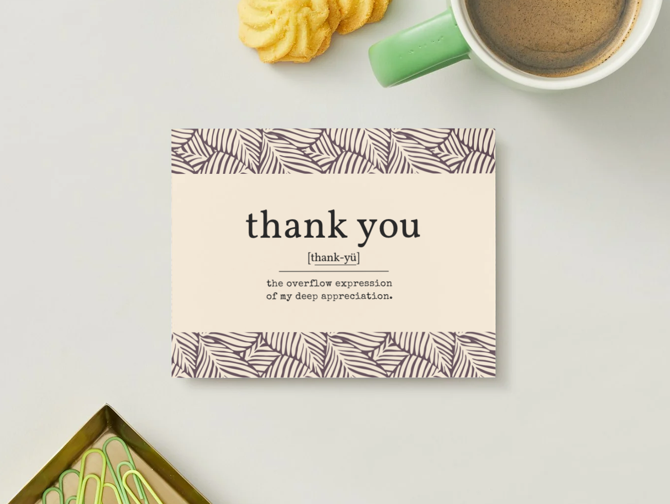 brown and beige thank you definition postcard with background leaves, up-close near green mug, paper clips, and two yellow cookies
