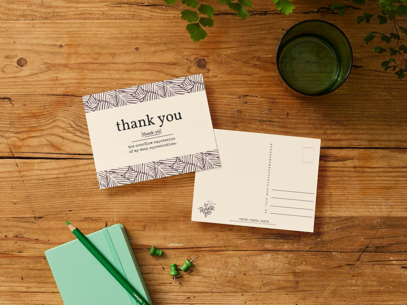 A flat-lay of a postcard and its back on a rustic wooden table, styled with a mint green notebook, a green pencil, pushpins, and a dark green glass cup.