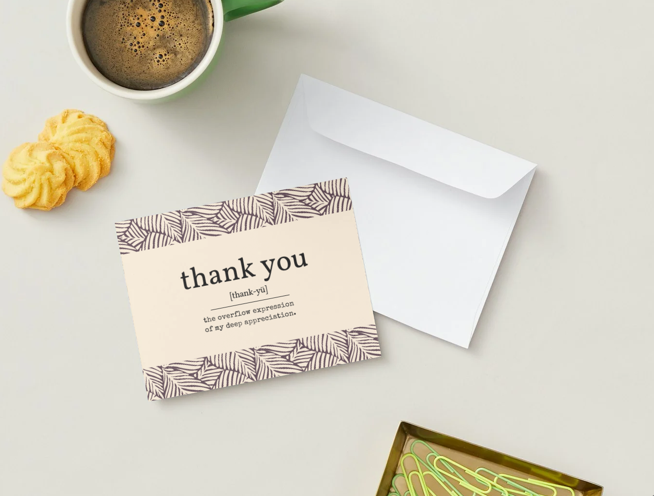 A styled flat-lay of a 'thank you' card with a white envelope, set on a light beige surface alongside a green coffee mug, two yellow cookies, and a box of green paperclips.
