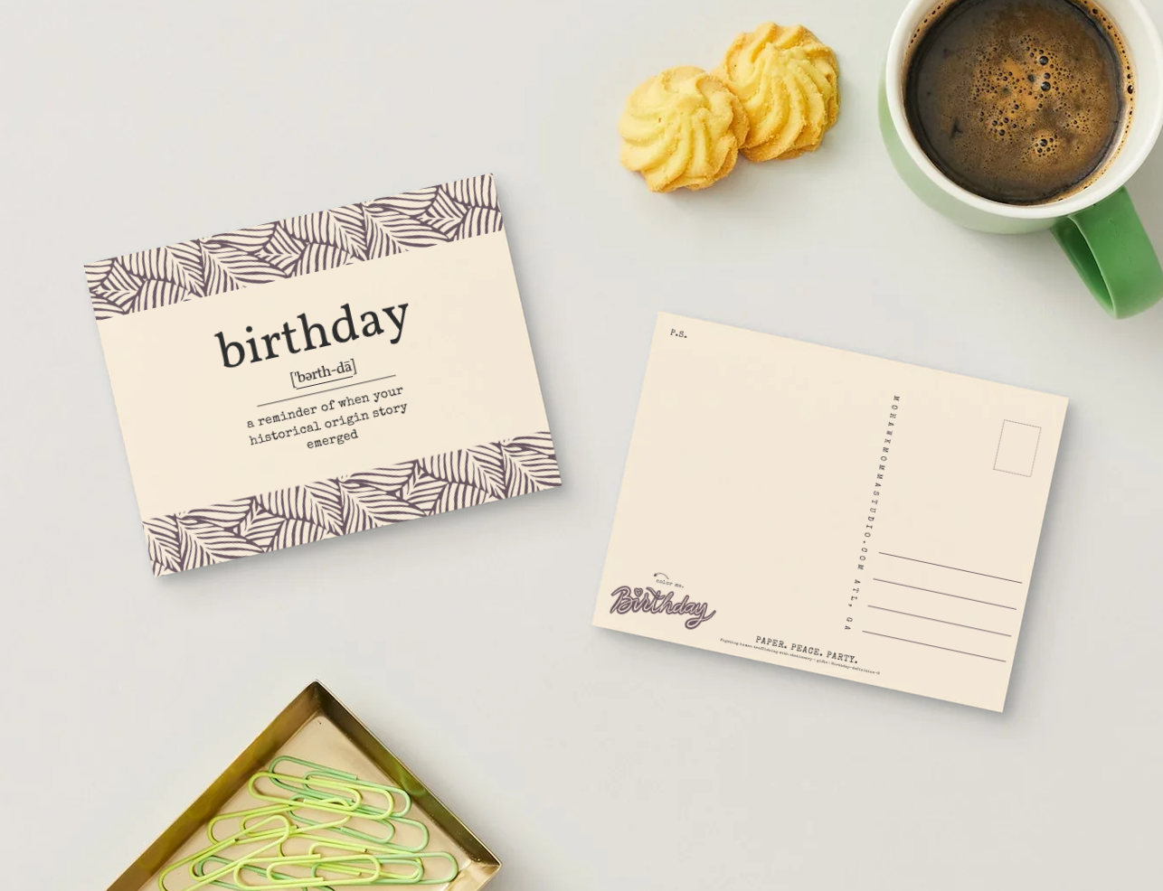 A styled flat-lay of a birthday postcard set on a light gray surface, with a front card reading 'birthday' and a matching back card, alongside cookies, a green mug of coffee, and paperclips.