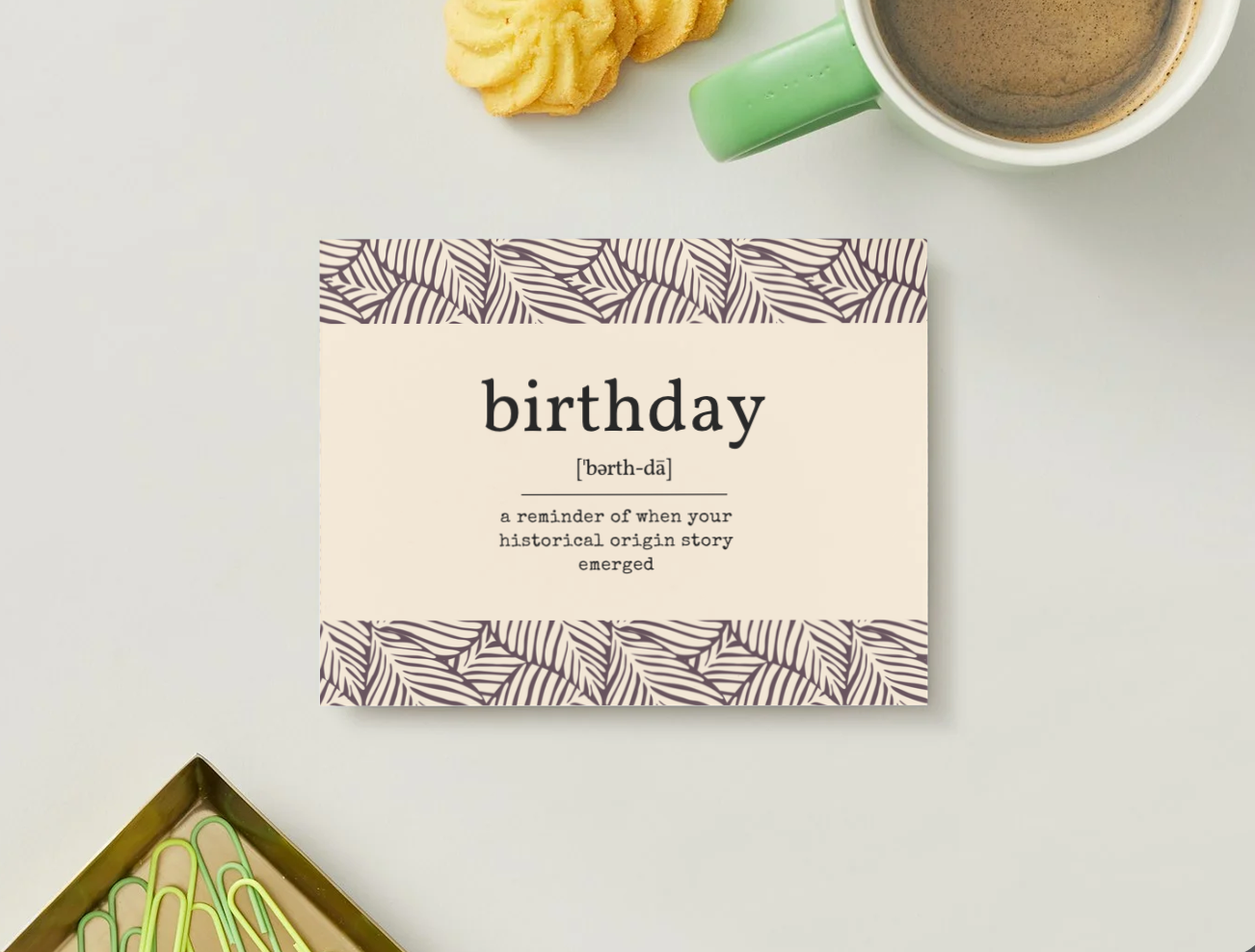 A square birthday card with a cream center and patterned borders sits on a light gray surface. The card reads 'birthday [birth-dá] a reminder of when your historical origin story emerged.