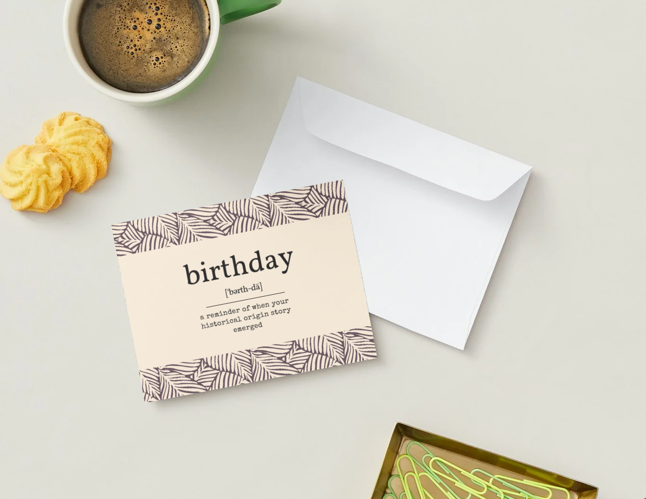 A greeting card with a patterned border and the word 'birthday' printed on it, shown on a light gray surface with an envelope, a green mug of coffee, and small cookies.