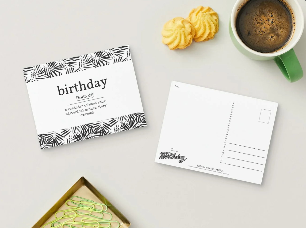 A styled flat-lay featuring a birthday postcard set on a light gray surface. The set includes a front card with a black palm leaf pattern border and the word 'birthday' in lowercase, a white postcard back with printed address lines and a decorative script logo, a green coffee mug filled with coffee, two yellow cookies, and a gold tray with green paperclips.