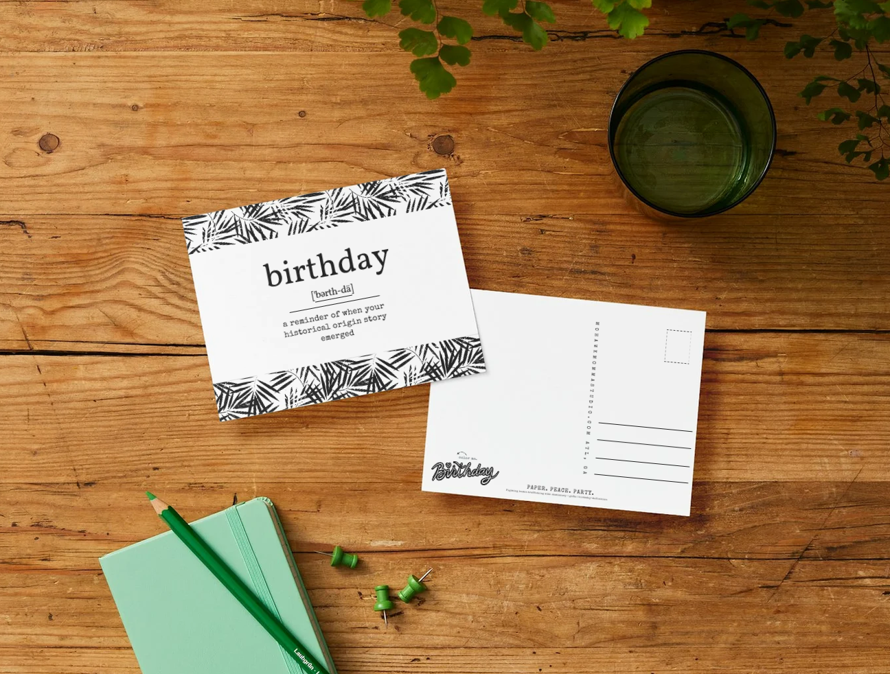 A flat-lay of a black and white tropical leaf patterned birthday postcard and its matching back on a rustic wooden table, styled with a mint green notebook, green pencil, and a dark green glass of water.