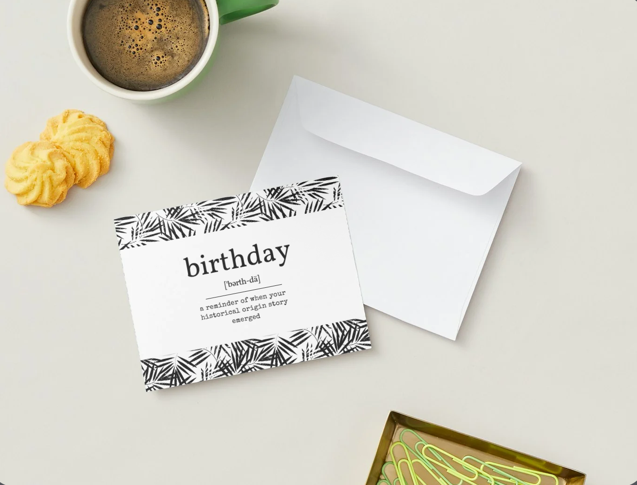 A styled flat-lay of a birthday greeting card and matching envelope on a light beige surface, with a green coffee mug, two yellow cookies, and a small decorative box partially visible.