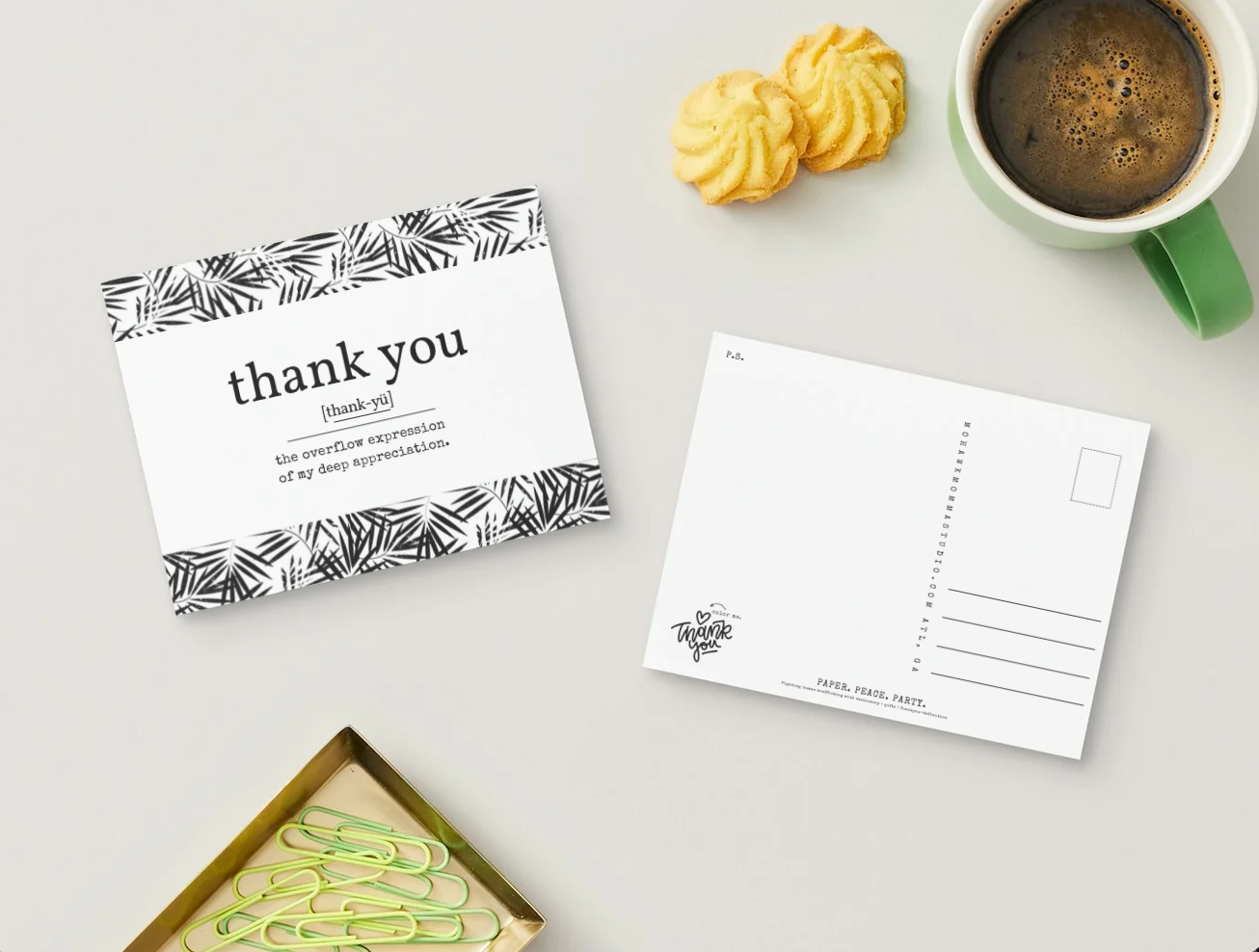 A styled flat-lay showing a printed thank-you card with black palm-patterned borders and a white postcard on a light gray surface, alongside a green mug of coffee, two yellow cookies, and paperclips in a box.