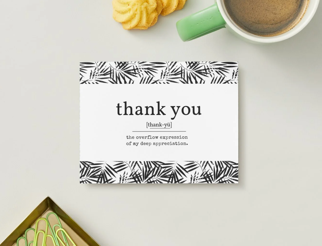 A square white thank-you card with black tropical leaf patterns at the top and bottom, styled on a light gray surface with a green mug, cookies, and a gold tray nearby.