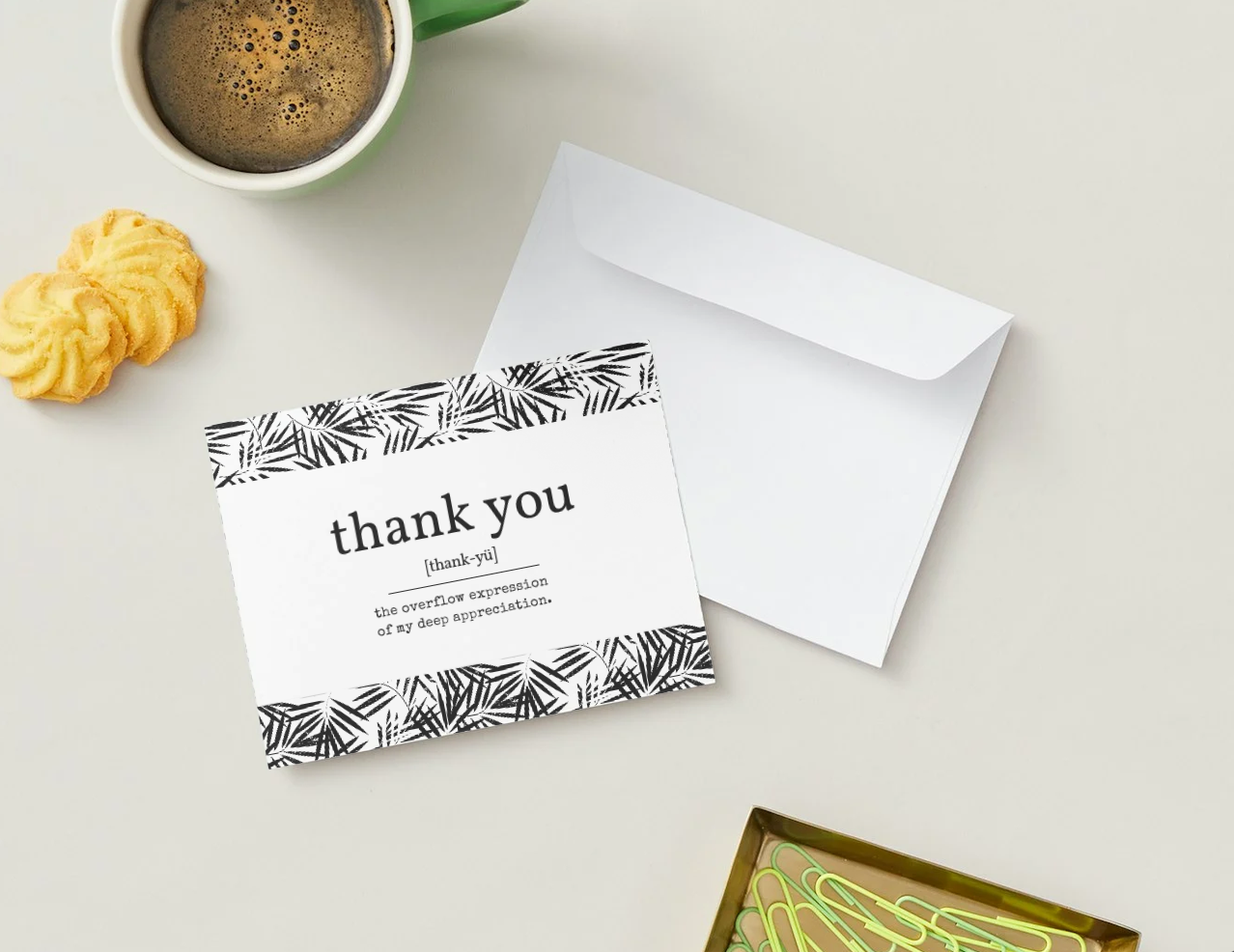 A styled flat-lay of a white thank you card with black palm-leaf patterned borders, placed next to a white envelope on a light beige surface. A green coffee mug, two yellow cookies, and a small box of paperclips are visible as props.