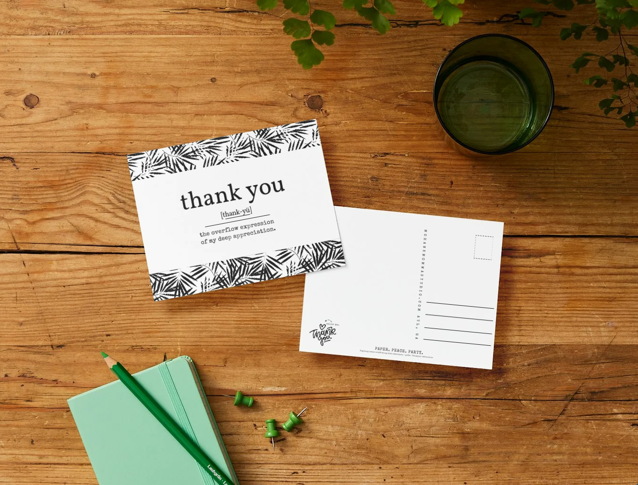 A flat-lay of a postcard set on a rustic wooden table, featuring a front card with a black palm leaf pattern and the text 'thank you' and an open back with writing lines. A mint green notebook, a green pencil, and a dark green glass cup are arranged nearby.